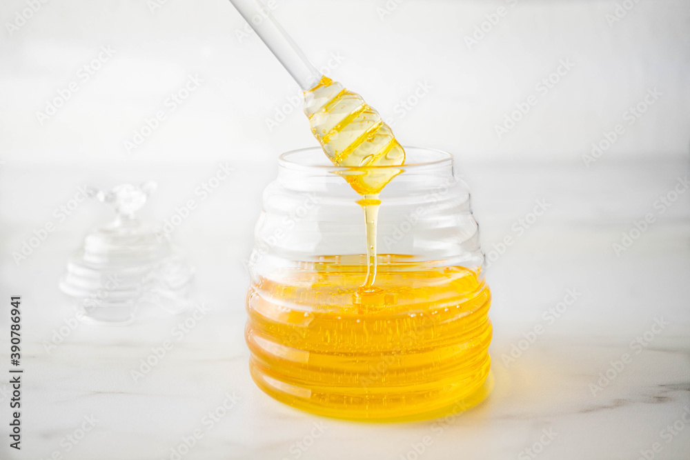 Honey flows from a honey spoon into a glass jar. Healthy organic thick honey in a transparent jar stands on a white table. Close-up
