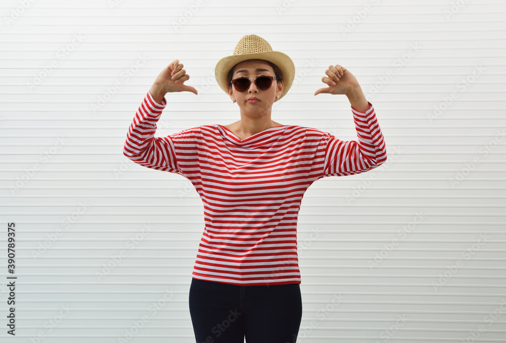 Naklejka premium Moody young asian woman wearing red stripped shirt, sunglasses, straw hat and jeans showing dislike thumps down hand sign standing over white wall background, Upset facial expression