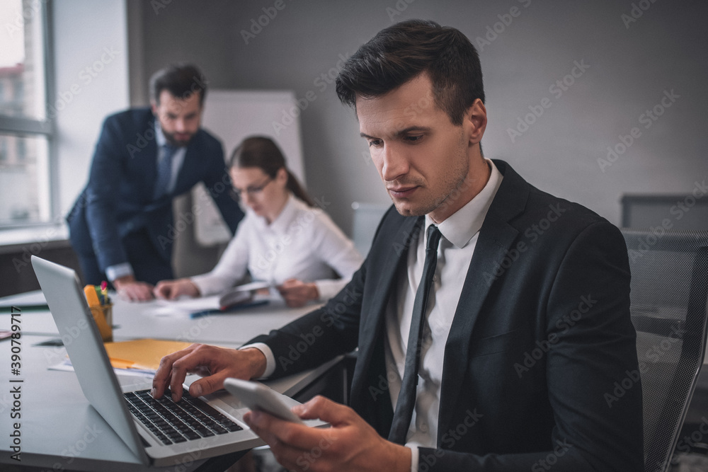 Man with laptop looking at smartphone in office Man with laptop looking at smartphone in office