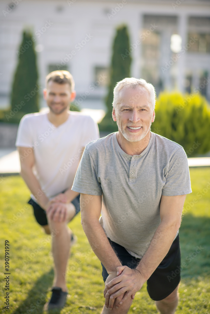 Young male and mature grey-haired male stretching legs outside, hands on knee, smiling