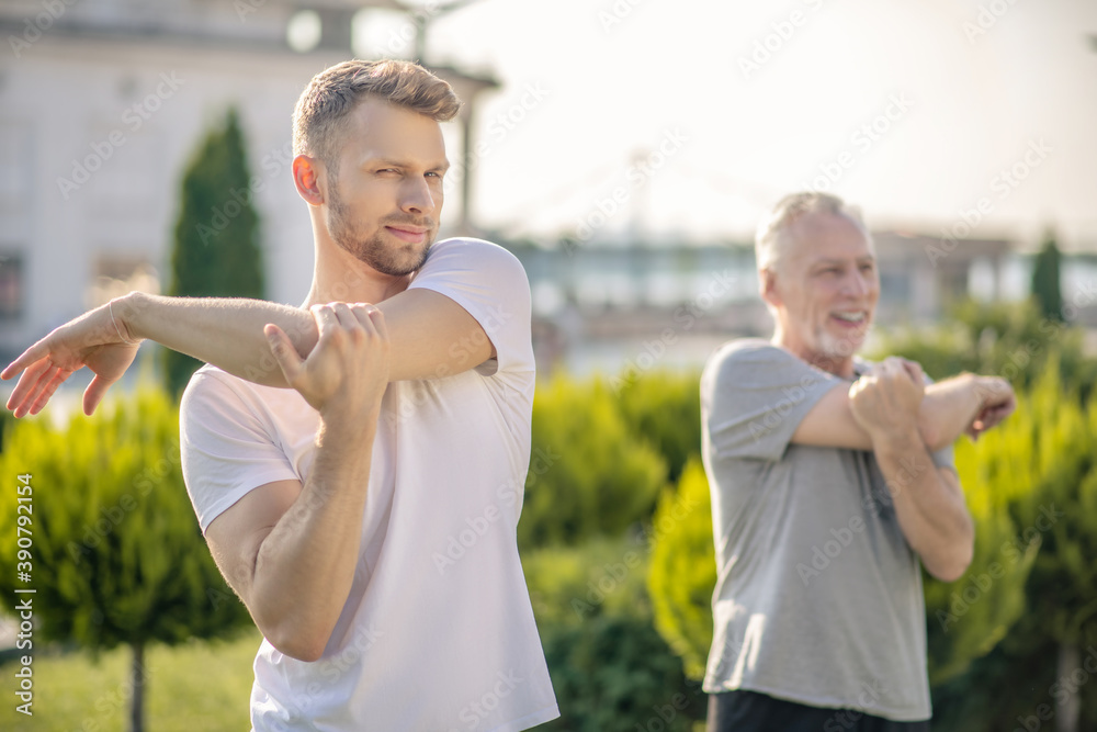 Obraz premium Young bearded male and mature grey-haired male stretching arms outside