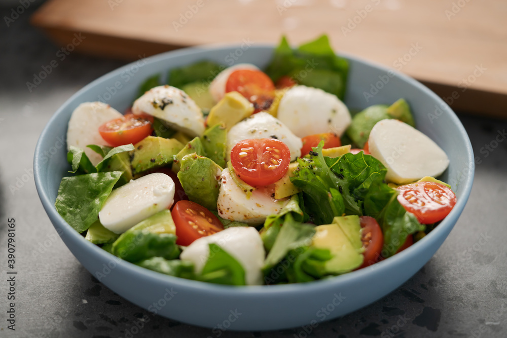 Salad with avocado, cherry tomatoes, romaine and mozzarella in blue bowl on concrete countertop