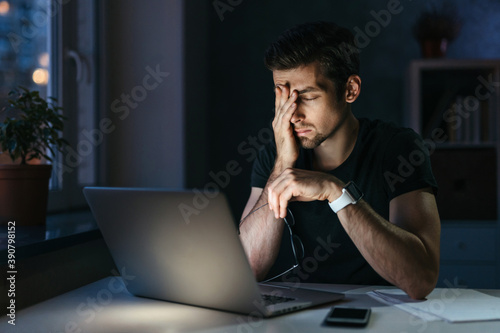 Young businessman has headache while working overtime with laptop at home office late night. Stressed depressed freelancer touching his head, feeling pain in eyes. Insomnia, internet addiction