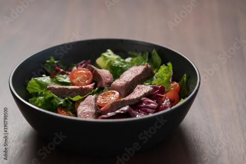 Steak salad with cherry tomatoes and mixed greens in black bowl on walnut wood background