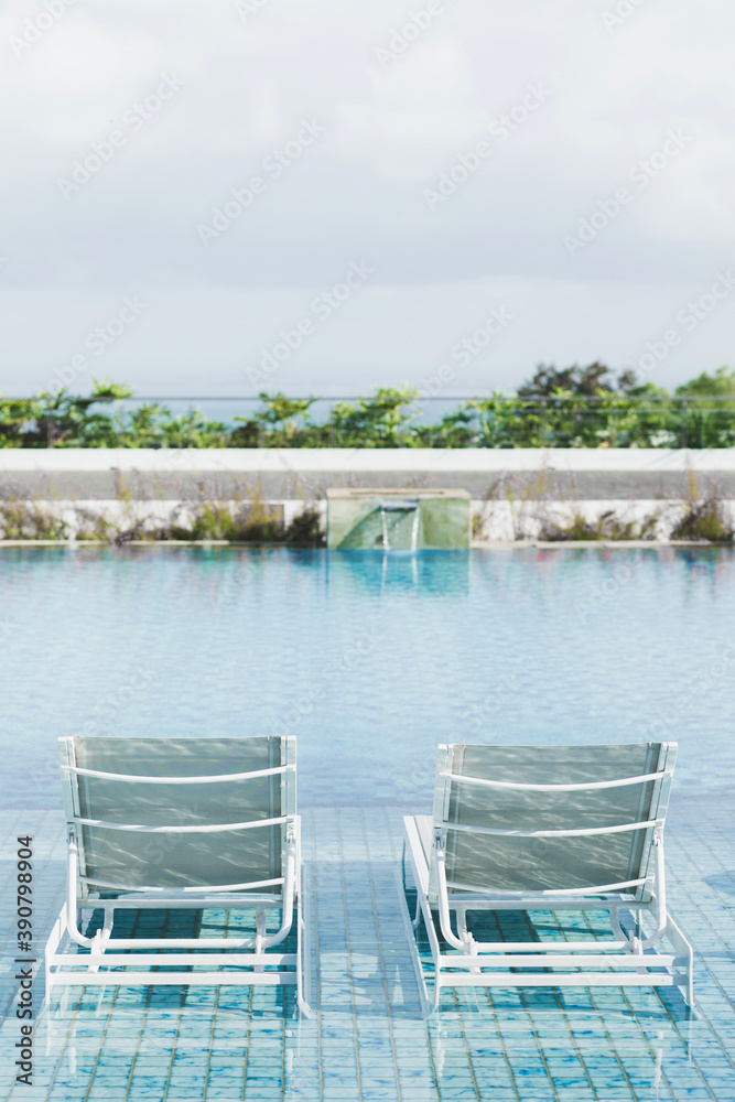 White metal recliners on pool side. Contemporary beach outdoor furniture. Swimming pool with clear blue water. Luxury vacations in Asia.