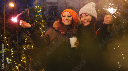 Two young smiling women holding burning sparkler and coffee in cold winter evening on street. Glowing blurred bokeh lights of Christmas