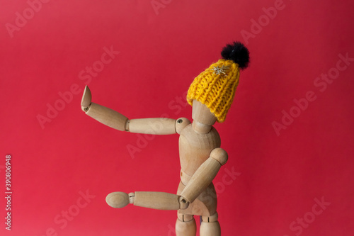 A wooden mannequin of a man in a knitted yellow hat points to a place next to him. Mannequin of a man made of wood on a red background.