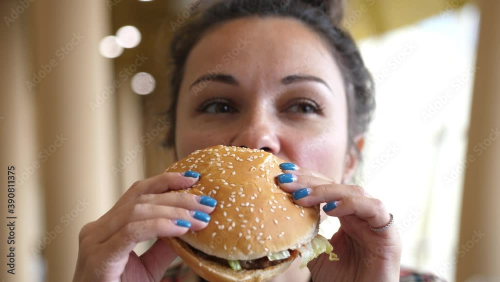 Young Hungry Woman Biting Vegan Meatless Burger in Fast Food Restaurant. Girl eating a hamburger. close-up shot. Fast food eats. Burger in female hands. Young Hungry Woman Biting Vegan Meatless Burger in Fast Food Restaurant. Girl eating a hamburger. close-up shot. Fast food eats. Burger in female hands.