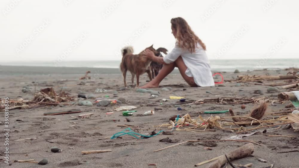 Full shot of young beautiful woman sitting on beach polluted with trash and plastic playing with two local dogs petting them with picturesque horizon in background