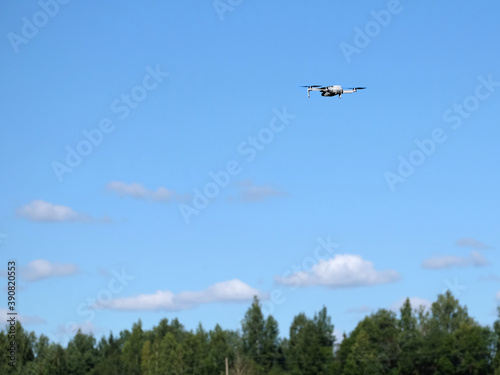 Quadcopter in flight above forest tree tops under blue sky with light white clouds in bright sunny summer day
