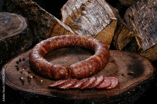 Ring of smoked sausage on a blackboard, on a background of burnt firewood. Several pieces of sausage sliced.
