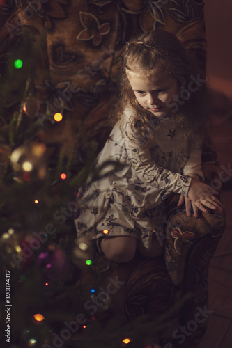 A cute little girl sits in an armchair in front of a decorated Christmas tree. Multi-colored bokeh. Christmas concept. Low key. Soft focus. Inside © Kiryakova Anna