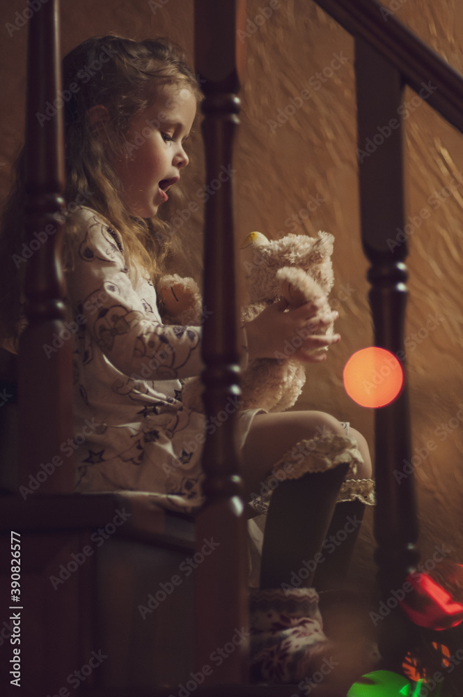 A cute little girl sits on a wooden ladder with a teddy bear in her hands and plays with him. Multi-colored bokeh. Christmas concept. Low key. Soft focus. Inside