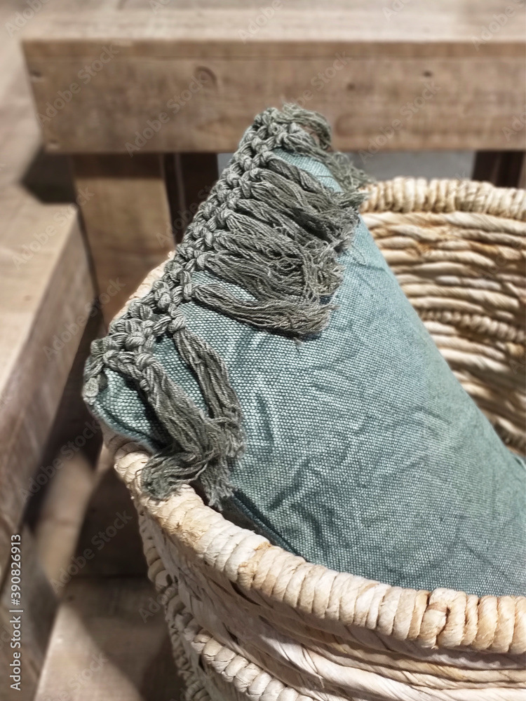 Fototapeta premium Basic vintage pale green cotton cushion with fringes in a wicker basket. Ethnic and hygge interior decoration. Corner detail of natural pillow with beaded fringe.