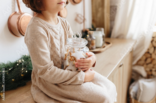 Toddler girl wearing beige dress holding glass jar with christmas cookies