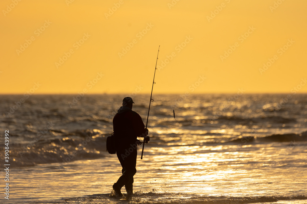 Silhouette of a fisherman walking through waves along the coast at sunset.
