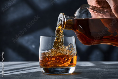 Round glass of whiskey with ice on a concrete table, a glass is filled with whiskey from a bottle.