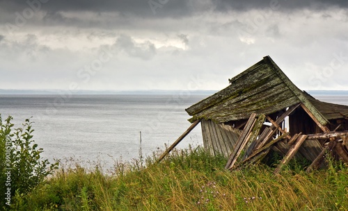 Old wooden ramshackle house on the bank the Lyokshmozero lake covered with grass against cloudy sky in summer in Arkhangelsk region, Russia.