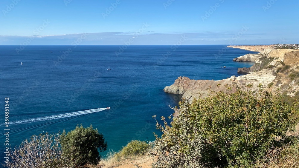 Panoramic seascape - calm azure sea, rocks and mountains surround the bay and a small boat in the distance in the fall on the Black Sea coast in Crimea, Cape Fiolent in Sevastopol.