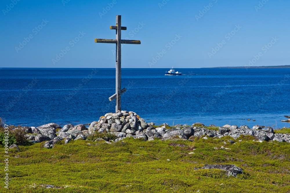 The wooden cross on a grave on the grassy shore of the White Sea and a ship in the background in summer on Solovetsky Islands, Russia.