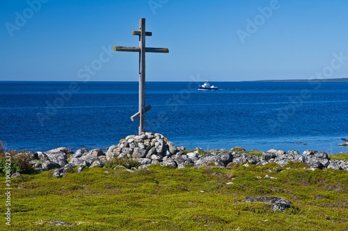 The wooden cross on a grave on the grassy shore of the White Sea and a ship in the background in summer on Solovetsky Islands, Russia.