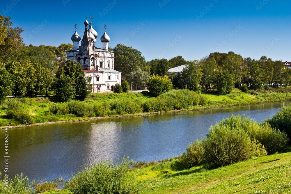 White orthodox Church of St. John of Chrysostom among green tall trees on the bank of the river in Vologda, Russia.