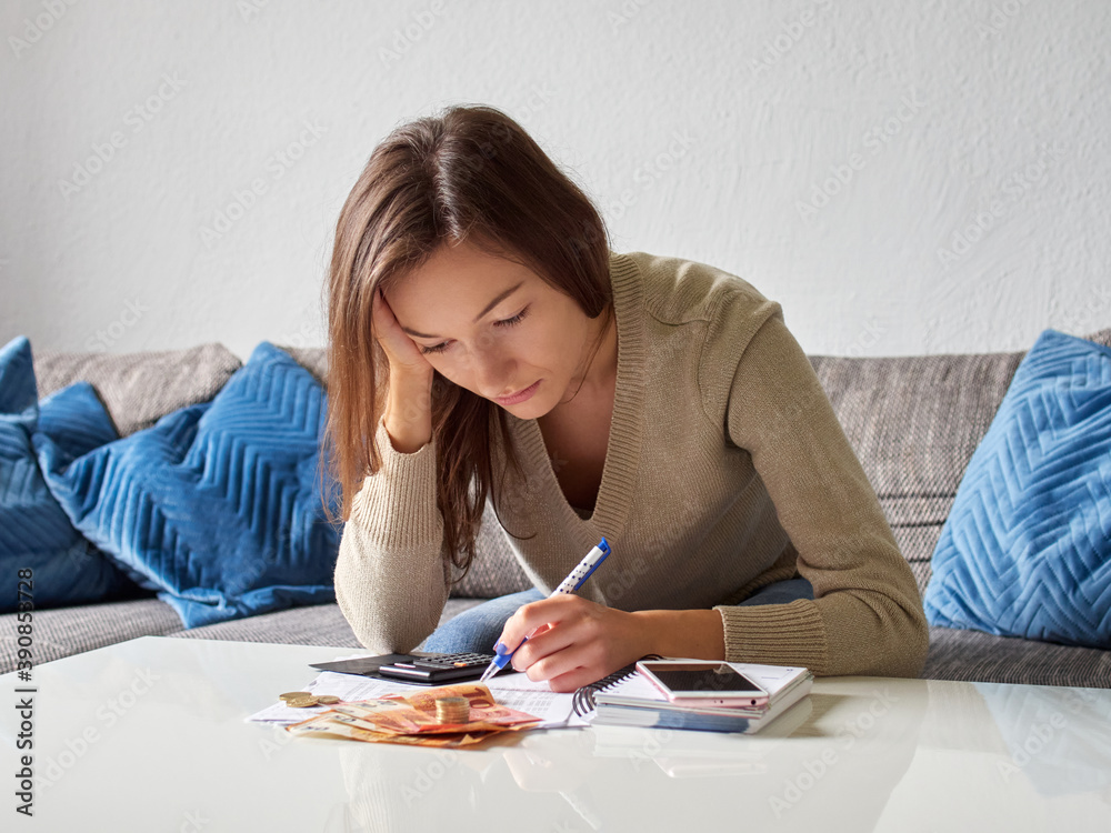 A young, depressed, stressed woman props her head with her hand. A pensive girl is counting bills in the living room. There is euro money and a calculator on the table. Summary of monthly costs. A young, depressed, stressed woman props her head with her hand. A pensive girl is counting bills in the living room. There is euro money and a calculator on the table. Summary of monthly costs.