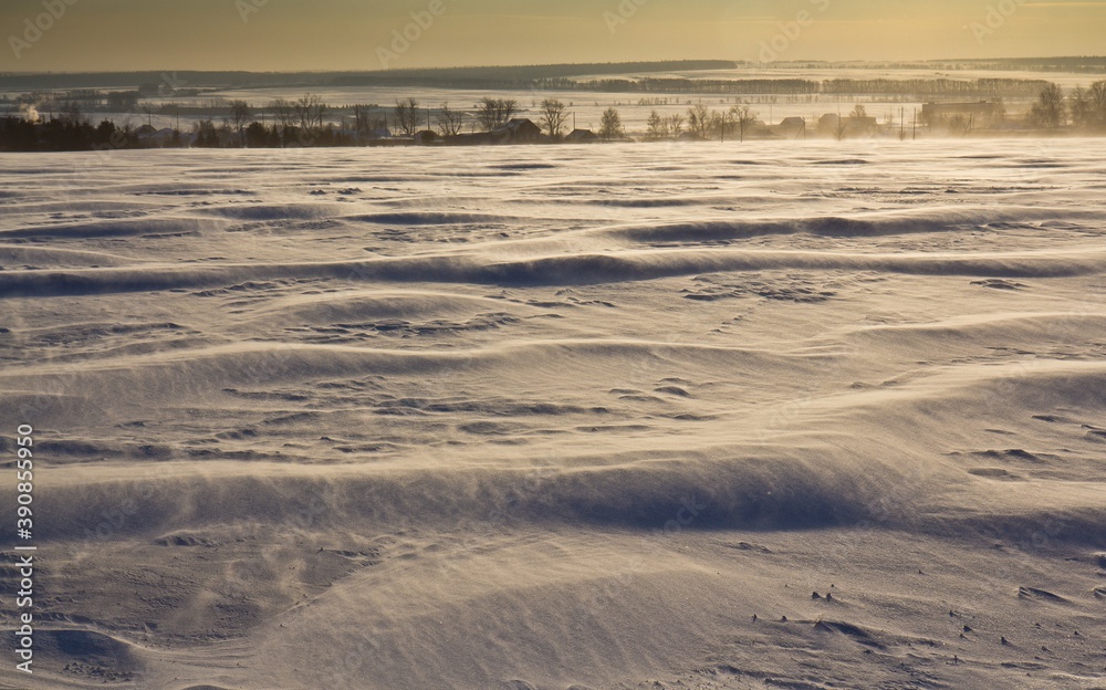 Snowy dunes (waves) in the frosty morning and small houses in the background in Udmurtia, Russia.