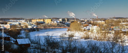The cityscape of russian city Kungur of Perm Krai: a bridge over the frozen river, bare trees on its bank and many houses and smoky chimneys in the background in snowy winter.