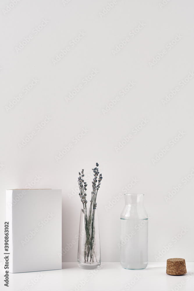 A transparent vase with lavender, a bottle of water with a cork stopper and a book on a white background. Minimalism, eco-materials in the interior decor. Copy space, mock up.
