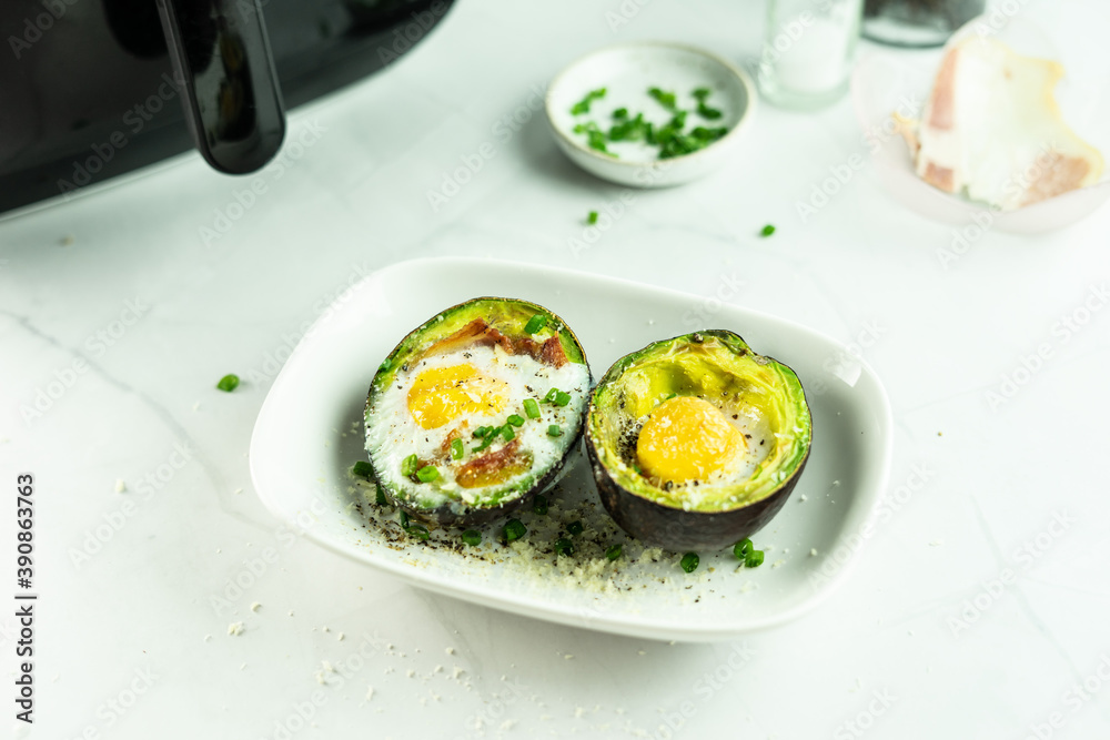 Baked eggs in avocado springkle with green onion and Parmesan cheese in a white dish, the Air Fryer and condiment behind, on the withe marble table. Healthy and keto breakfast. Eggs disk..