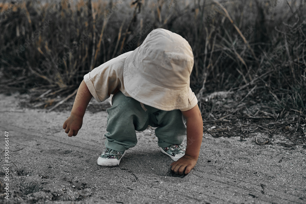 A two-year-old child in a hat crouched touches something on a country road, summer. Experience of the world.