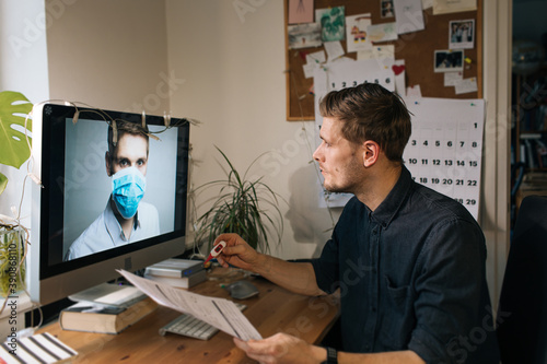 Caucasian man wearing casual shirt using computer for telemedicine. Online doctors in video call. Telehealth during pandemic. Video consult in the computer monitor doctor wear medical face mask