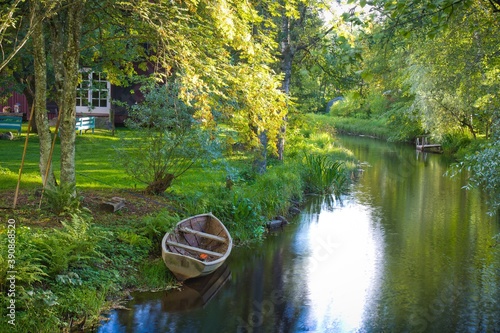 Pictursque rural view of small calm river flowing among green forest trees, and boat on its bank. Nice wooden house is seen behind the trees. Finland. photo