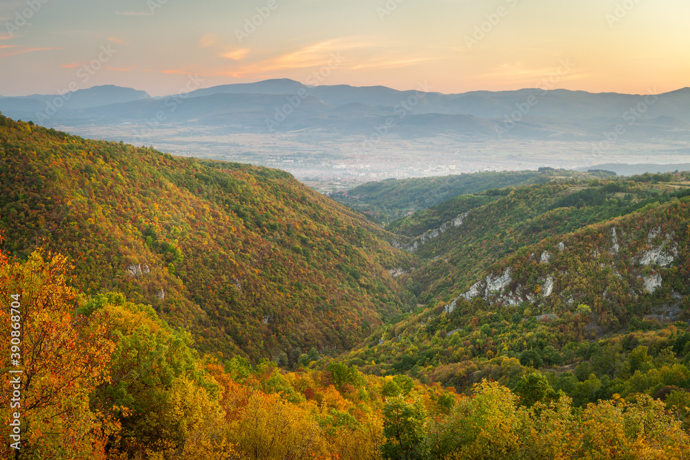 Autumn colored trees around the rocky canyon of Gradasnica river, leading towards misty, hazy Pirot cityscape and colorful blue hour sky