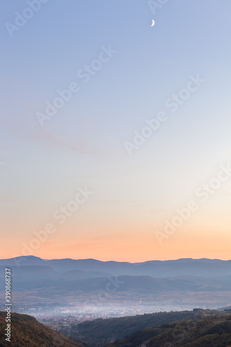 Soft, misty view of distant Pirot city, foreground hills covered by autumn colored trees and colorful sunset sky with young moon