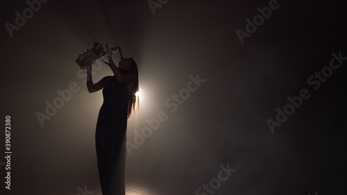 Young beautiful girl in a dark dress plays on a golden shiny saxophone on stage. Dark studio with smoke and stage lighting. Hands and saxophone close up. Side view.Slow motion video
