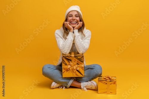 young smiling happy pretty woman sitting with golden present boxes celebrating new year, christmas gifts, wearing white knitted sweater, scarf and hat, winter fashion trend, on yellow background © mary_markevich