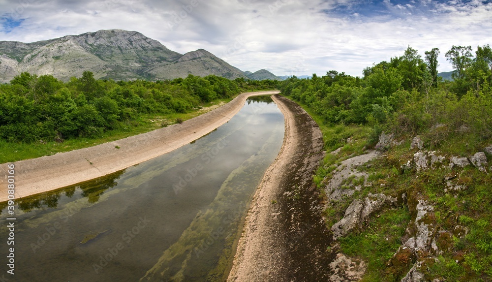 The artificial channel with concrete embankment surrounded with green bushes and mountains in the background against cloudy sky in Bosnia and Herzegovina.