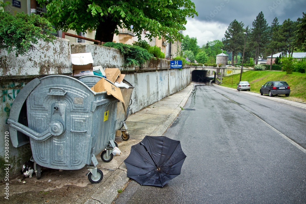 A road going under the tunnel surrounded with green trees with parked cars and a dumpster full of carton boxes and a black opened umbrella lying around in Cetinje, Montenegro. A road going under the tunnel surrounded with green trees with parked cars and a dumpster full of carton boxes and a black opened umbrella lying around in Cetinje, Montenegro.