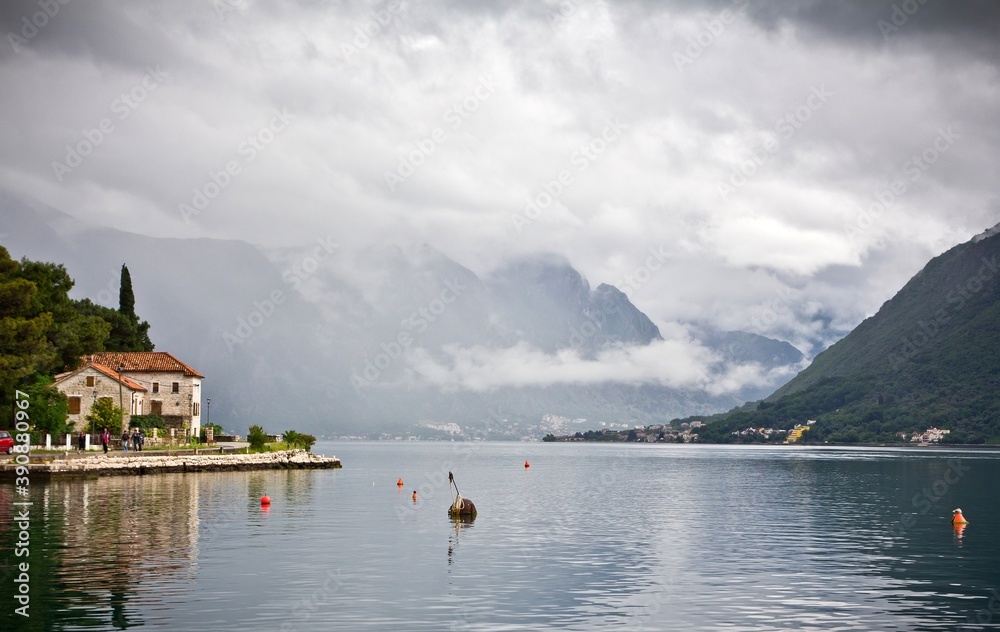 The stone embankment of the Bay of Kotor with buoys, small tile roof houses and mountains in clouds in the background in Perast, Montenegro. The stone embankment of the Bay of Kotor with buoys, small tile roof houses and mountains in clouds in the background in Perast, Montenegro.
