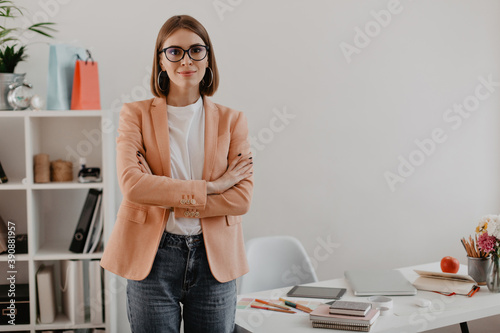 Female entrepreneur in excellent mood looking at camera with her arms crossed against bright workplace