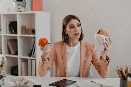 Picture of business woman in pink jacket sitting in workplace. Woman choose between tasty burger and wholesome apple