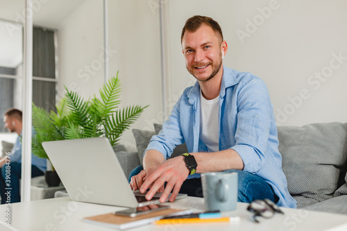 handsome smiling man in shirt sitting relaxed on sofa at home at table working online on laptop from home freelancer, social distancing isolation communication digital conference, looking in camera