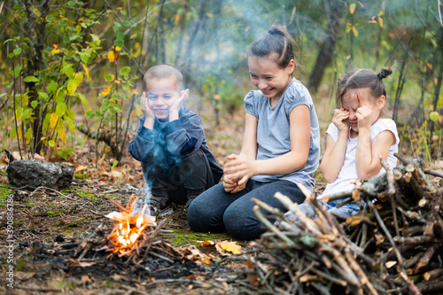 Children sitting near campfire in forest and closing eyes because of smoke