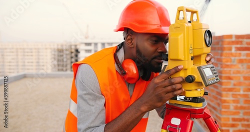 African American young man topographer in casque measuring angle with total station on roof of building. Male builder. Constructor doing topographic measures. Geodesy concept. Geodesy. Constructing. photo