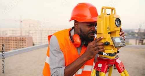 African American young man topographer in casque measuring angle with total station on roof of building. Male builder. Constructor doing topographic measures. Geodesy concept. Geodesy. Constructing. photo