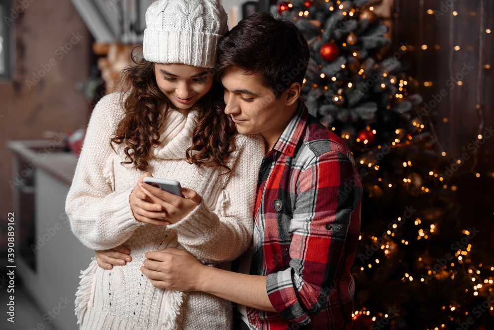 Happy young couple checking Christmas messages on a smartphone hugging standing near decorated christmas tree enjoying spending time together. Joyful cozy moments in winter holidays. Advertising