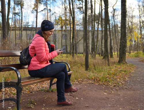 A woman sits on a bench in an autumn park, drinks hot coffee and looks into the smartphone screen