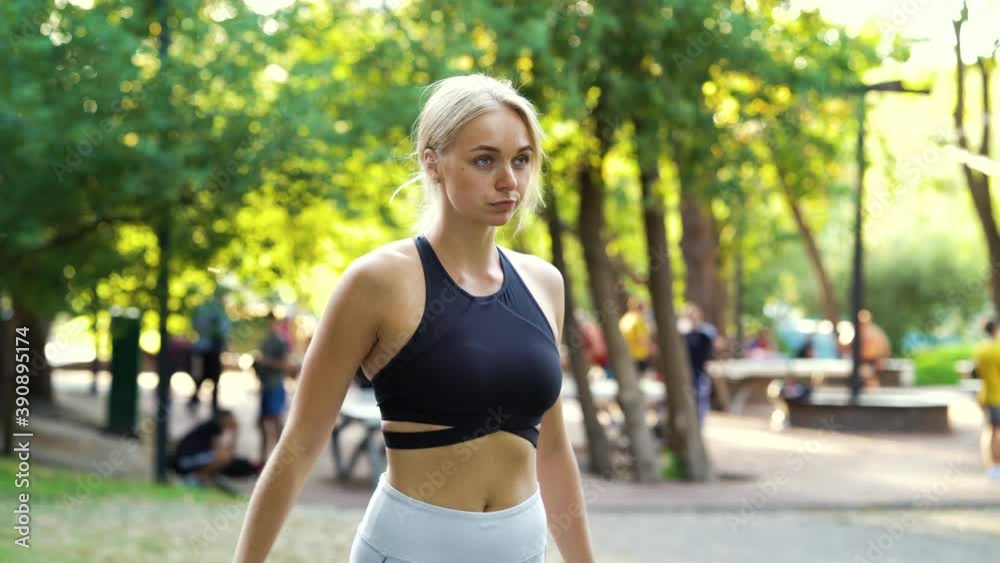Blonde woman wearing sports top standing in sunny park and preparing to start, blurred people on background. Female jogger training outside. Concept of sport